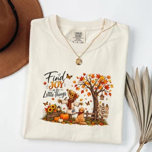 May include: A cream-colored t-shirt featuring a graphic of a scarecrow, a dog, pumpkins, sunflowers, and a tree with autumn leaves. The text reads Find JOY in the Little Things and Breathe Be Grateful Stay Kind. A gold necklace rests on the collar.