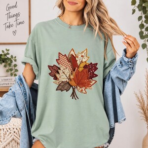 Puede incluir: Una camiseta de cuello redondo color verde salvia con un gráfico central de un conjunto de hojas otoñales en varios patrones de tela, incluyendo cuadros, flores y tonos sólidos marrones, naranjas y amarillos, sobre una camisa vaquera azul.