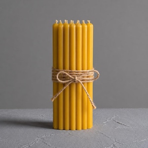 Puede incluir: Un conjunto de velas amarillas de cera de abeja atadas con hilo de cáñamo. Las velas son cilíndricas y tienen una superficie ligeramente texturizada. El hilo está envuelto alrededor del centro de las velas y atado con un lazo. El fondo es gris liso.