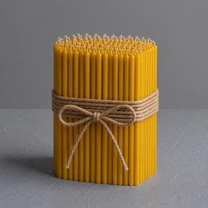 May include: A bundle of yellow beeswax candles tied with a natural twine bow. The candles are cylindrical and arranged in a rectangular shape. The background is a solid gray.