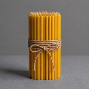 Puede incluir: Un paquete de velas amarillas de cera de abeja atadas con hilo natural. Las velas son cilíndricas y tienen una superficie ligeramente texturizada. El hilo está atado con un lazo en el centro.