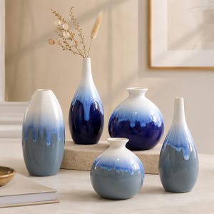 May include: A set of five ceramic vases in varying shapes, featuring a white to blue and grey gradient design. The vases range from tall and thin to round. One vase holds dried flowers. The vases are suitable for home decor.
