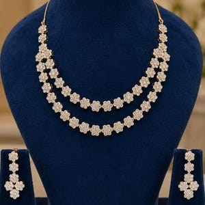 以下が含まれることがあります： 青いベルベットのバストに飾られた、ゴールドトーンのネックレスとイヤリングのセット。ネックレスは、花型の装飾が2連になっています。お揃いのイヤリングも花型です。ジュエリーは、柔らかくぼやけた背景を背景にしています。