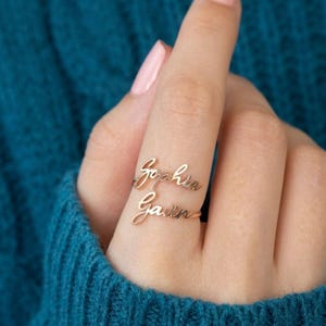 Peut inclure: Une bague dorée à deux rangs, chacune portant un nom en écriture cursive. La rangée supérieure indique "Sophia", et la rangée inférieure indique "Gavin". La bague est portée au doigt, sur un fond bleu sarcelle.