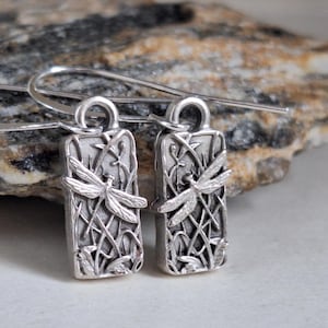 May include: A pair of silver-toned rectangular drop earrings featuring a raised dragonfly motif set against a background of textured reeds. The earrings are attached to simple silver-toned ear wires, showcasing a metallic finish with dark oxidized detailing.