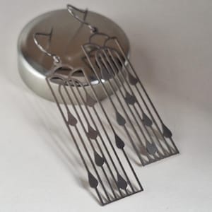 May include: A pair of silver-coloured, geometric dangle earrings. Each earring has a rectangular design with vertical lines and teardrop-shaped cutouts. The earrings are displayed on a silver lid against a white background.