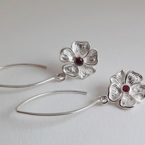 May include: A pair of silver-colored flower earrings with a small, round, red gemstone in the center of each flower. The earrings have a hook design and a curved, elongated shape.