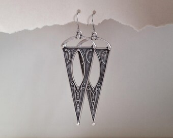 May include: A pair of silver-toned earrings with a triangular design. Each earring features an oval cutout in the center and is adorned with intricate dot and line patterns. The earrings hang from simple hooks.
