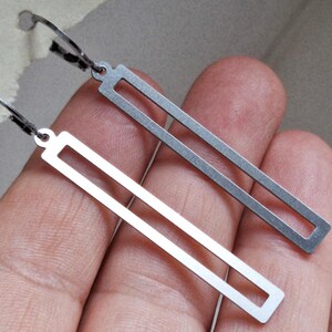 Op de afbeelding: Twee zilverkleurige rechthoekige oorbellen met een minimalistisch ontwerp. Elke oorbel heeft een lange, open rechthoekige vorm die aan een kleine haak hangt. De oorbellen worden tegen een neutrale achtergrond getoond.