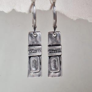 Może przedstawiać: Para srebrnych, prostokątnych kolczyków wiszących. Każdy kolczyk ma teksturowaną powierzchnię z centralnym prostokątnym wzorem i paskiem z koralików. Kolczyki mają zapięcie dźwigniowe.