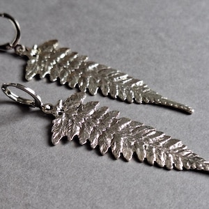 May include: A pair of silver-colored fern leaf earrings. Each earring features a detailed, textured leaf design with a lever-back closure. The earrings are displayed against a gray background, highlighting their intricate details.