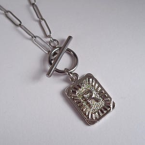 Op de afbeelding: Zilverkleurige ketting met een rechthoekige hanger met de letter "R". De hanger heeft een textuurontwerp en hangt aan een tuimelsluiting. De ketting is gemaakt van langwerpige schakels.