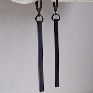 May include: A pair of black, minimalist earrings. Each earring features a small hoop that connects to a long, rectangular bar. The earrings are suspended against a neutral background, highlighting their sleek, modern design. These earrings are a simple yet elegant accessory.