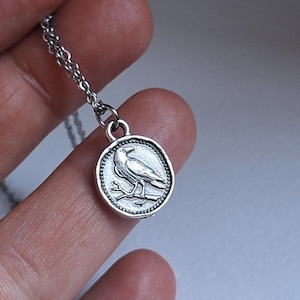 Op de afbeelding: Een zilverkleurige metalen ketting met een ronde hanger met een reliëf van een vogel op een tak. De hanger heeft een rand met kraaltjes en hangt aan een fijne zilverkleurige metalen schakelketting.
