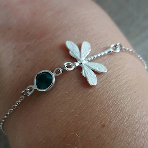 Könnte beinhalten: Ein silbernes Armband mit einem Libellenanhänger und einem dunkelgrünen Edelstein. Die Libelle hat detaillierte Flügel und ist an einer feinen Kette befestigt. Der Edelstein ist oval.