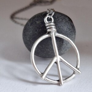 May include: A silver peace symbol pendant necklace. The pendant, a classic peace sign, hangs from a fine chain. The necklace is set against a dark grey stone, with a soft, neutral background.