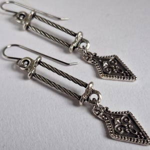 May include: A pair of silver-toned drop earrings featuring two parallel twisted metal cables connecting a hook wire to a diamond-shaped pendant. The pendant has an oxidised finish with embossed scrollwork and a beaded border detail.