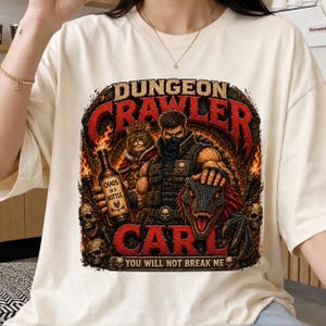 Puede incluir: Una camiseta color crema con un estampado gráfico que incluye el texto "DUNGEON CRAWLER CARL" y "YOU WILL NOT BREAK ME". El diseño muestra un personaje enmascarado, un gato, una cabeza de dinosaurio, calaveras y una botella "CHAOS IN A BOTTLE".