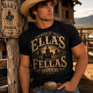 May include: A black short-sleeved t-shirt featuring a graphic print with the text 'MEMBER OF THE ELLA'S FELLAS CLUB' in gold lettering, surrounding an illustration of a cowboy on horseback in a desert landscape at sunset.