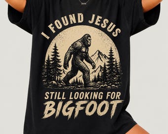 Pode incluir: Uma t-shirt preta de manga curta com uma estampa gráfica de uma silhueta de Bigfoot a caminhar por uma floresta com montanhas ao fundo. O texto diz I FOUND JESUS STILL LOOKING FOR BIGFOOT num estilo de letra bege desgastado.