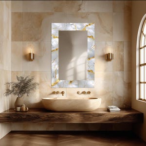 May include: A rectangular wall mirror featuring a white marble frame with prominent gold veining. The mirror is mounted vertically above a beige oval vessel sink and a dark wood countertop, reflecting the surrounding light and wall space.