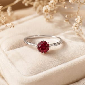 Pode incluir: Um anel de tom prateado com uma pedra preciosa vermelha profunda, redonda e facetada, montada numa cravação de seis garras. A banda é fina e polida, apresentando um design simples que destaca a cor vibrante e a clareza da pedra central.