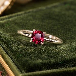 Op de afbeelding: Een ring van gepolijst zilverkleurig metaal met een enkele dieprode edelsteen in kussenslijpvorm, gezet in een zetting met vier pootjes. De ringband is glad en slank en toont een levendige, verzadigde rode steen in het midden van het ontwerp.