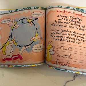 May include: An open children's book with two pages showing illustrations of yellow fireflies and a person. The left page shows fireflies flying in a circle with speech bubbles saying outstanding, outrageous, and out of this world. The right page has the text The Story of Small o and practice lines for cursive writing.