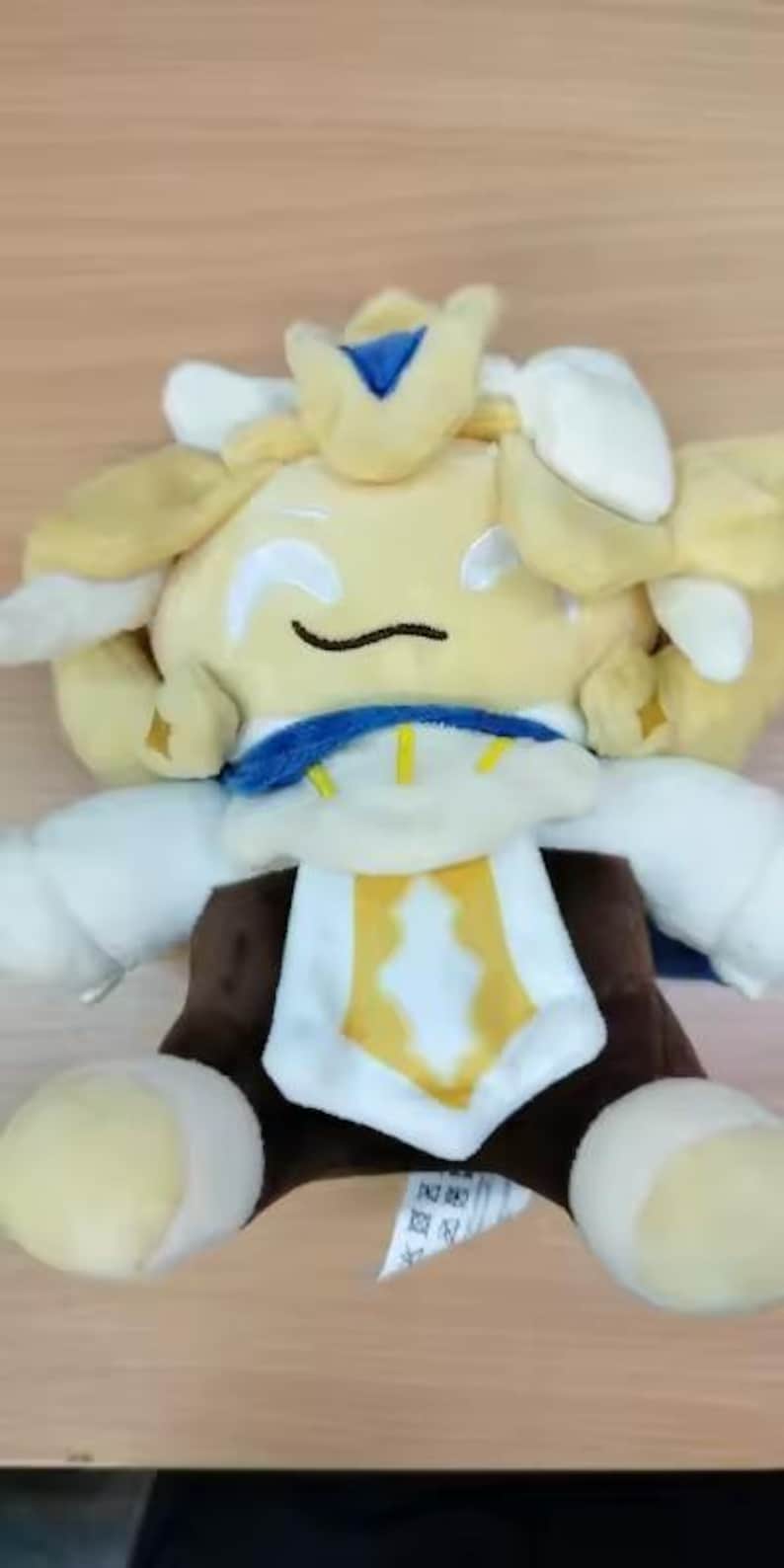 May include: A plush toy featuring a character with pale yellow hair, white and blue accents, and a brown body. The character has embroidered white eyes and a curved black mouth. A white and yellow chest piece is centered on the torso.