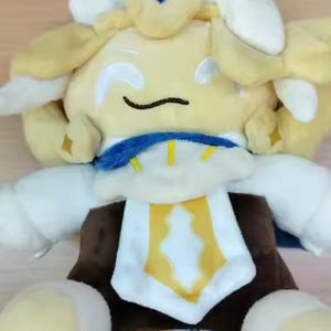 May include: A plush toy featuring a character with light yellow hair, white and blue accents, and a brown body. The character has embroidered closed eyes and a curved mouth, wearing a white collar with blue trim and a central yellow emblem.