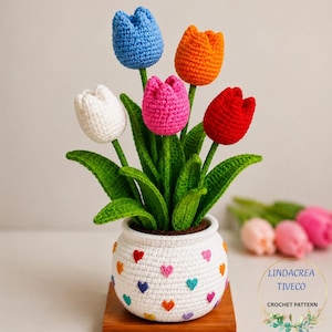 May include: A crocheted tulip arrangement in a white pot decorated with colorful hearts. The arrangement features five tulips in blue, orange, red, pink, and white. The green leaves and stems add a natural touch to this handmade craft.