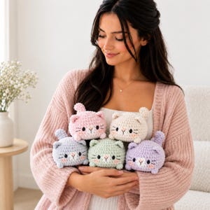 May include: A collection of five small, round, crocheted cat plushies in pastel colors including pink, cream, light blue, mint green, and lavender. Each cat features embroidered black eyes, a pink nose, and whiskers, with a small tail.