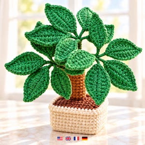 May include: A crocheted potted plant featuring a brown trunk and numerous vibrant green leaves. The plant is set in a square, cream-colored crocheted pot with a dark brown top surface, showcasing detailed yarn stitching throughout the entire structure.