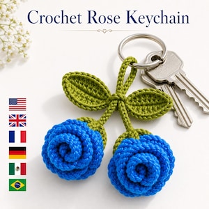 May include: A crochet rose keychain featuring two vibrant blue yarn roses attached to green crocheted stems and leaves, connected to a silver metal keyring with two attached keys. The text Crochet Rose Keychain is visible at the top.