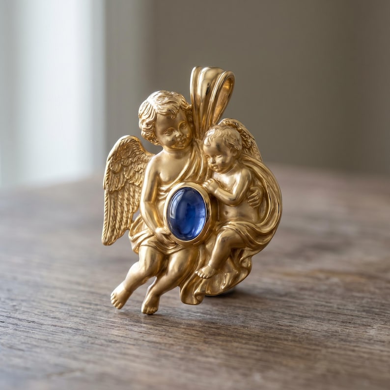 May include: A gold-colored pendant featuring two cherubic figures with wings, holding a large, oval, blue gemstone. The pendant has intricate details and a loop for a chain.