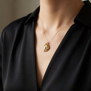 May include: A gold-colored pendant necklace featuring a detailed, ornate design with a small, dark blue gemstone at its center. The pendant hangs from a delicate gold chain. The necklace is displayed against a black top.