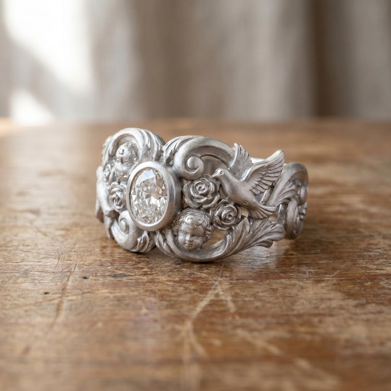 Puede incluir: Un anillo plateado con una gema clara de corte ovalado en el centro. El anillo presenta detallados relieves de rosas, querubines y una paloma. El anillo se exhibe sobre una superficie de madera.