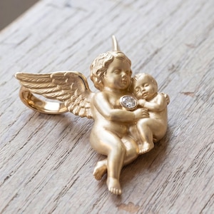 May include: A gold-colored ring featuring a cherub holding a baby, with a diamond accent. The ring has detailed wings and is set against a wooden surface. The ring's design is a representation of love and protection.