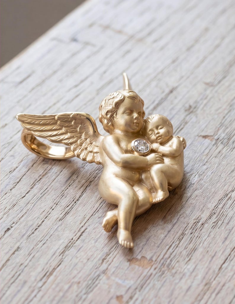 May include: A gold-colored ring featuring a cherub holding a baby, with a diamond accent. The ring has detailed wings and is set against a wooden surface. The ring's design is a representation of love and protection.