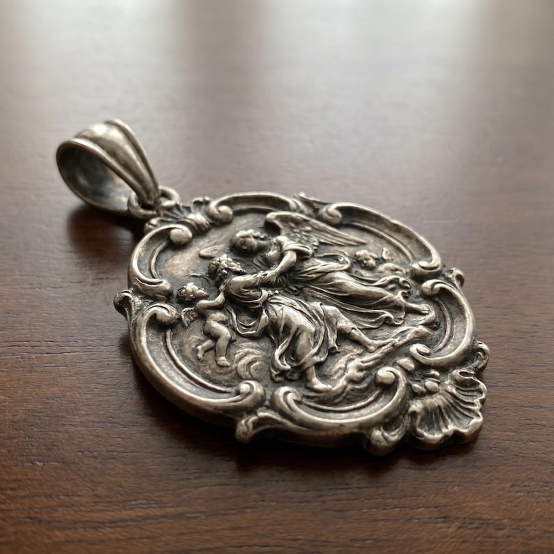 May include: A silver-toned metal pendant featuring an oval shape with an ornate, scrolled border. The center depicts a relief scene of a winged figure interacting with a smaller figure, surrounded by decorative flourishes and a large top bail.