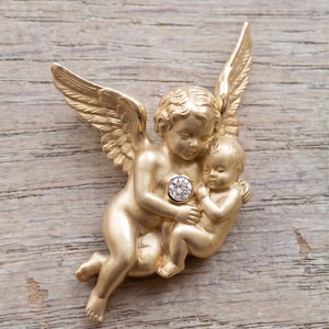 May include: A gold-colored cherub brooch featuring an angel holding a baby, with a sparkling gemstone at the center. The angel has detailed wings and is sculpted in a classical style. The brooch is set against a wooden background.