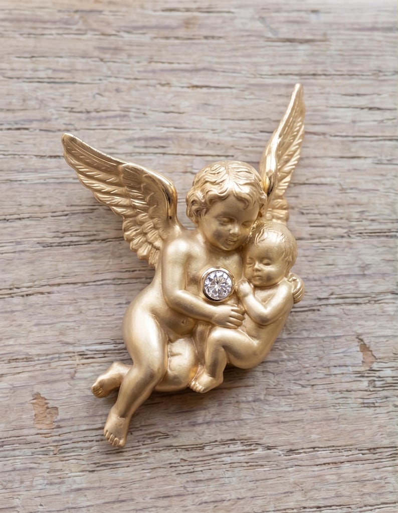 May include: A gold-colored cherub brooch featuring an angel holding a baby, with a sparkling gemstone at the center. The angel has detailed wings and is sculpted in a classical style. The brooch is set against a wooden background.