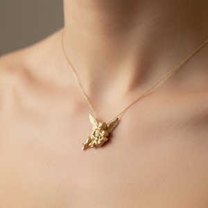 May include: A gold-colored necklace featuring a pendant of two cherubs embracing, with small wings and a small diamond. The pendant hangs from a delicate gold chain. The necklace is worn on a person's neck.