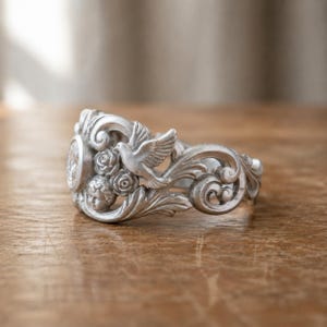 Puede incluir: Un brazalete plateado con detalles ornamentados. El dise&ntilde;o incluye una paloma, rosas y caras de querubines. El brazalete tiene una est&eacute;tica vintage, con patrones arremolinados y un relieve detallado.