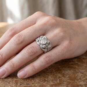 Puede incluir: Anillo plateado ornamentado con una gema transparente central, rodeado de intrincados dise&ntilde;os florales y de volutas. Las intrincadas tallas del anillo y la brillante gema crean un contraste llamativo, que sugiere un estilo vintage. El anillo se muestra en una mano.