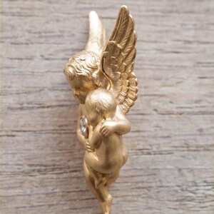 May include: A gold-colored cherub figurine with detailed wings, holding a smaller cherub. A clear gemstone is set in the chest of the larger cherub. The figurine is set against a light brown wooden background.