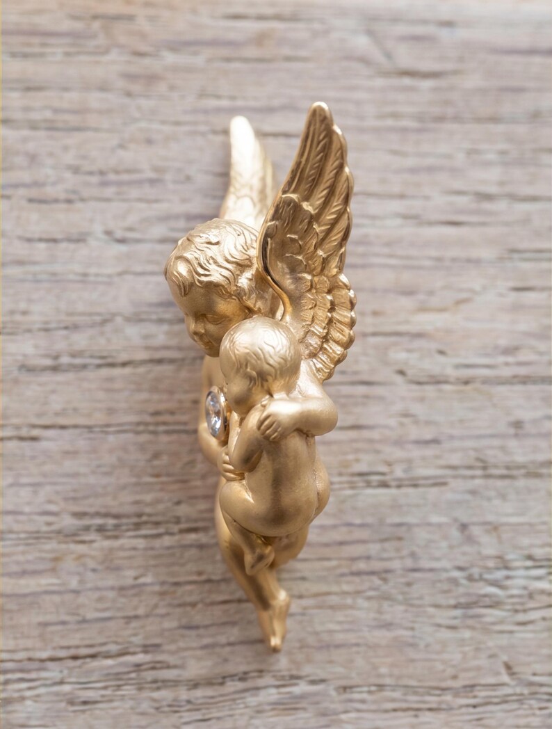 May include: A gold-colored cherub figurine with detailed wings, holding a smaller cherub. A clear gemstone is set in the chest of the larger cherub. The figurine is set against a light brown wooden background.