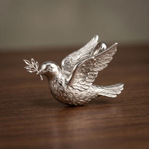 Op de afbeelding: Zilveren duifbeeldje met uitgespreide vleugels, een olijftak in de snavel. De vogel heeft gedetailleerde veren en een gepolijste afwerking. De duif, een symbool van vrede, staat tegen een donkere houten achtergrond.