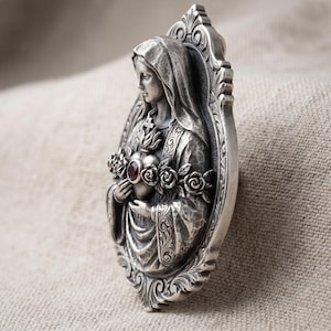 Puede incluir: Un colgante ovalado de tono plateado con una escultura en relieve de una figura con túnica y capucha. La figura sostiene un corazón adornado con una pequeña piedra preciosa roja circular en el centro, rodeado por una guirnalda de rosas esculpidas.