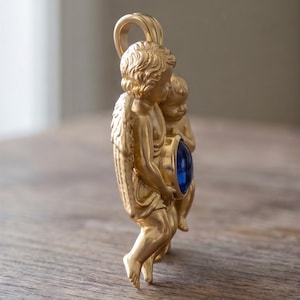 May include: A gold-colored pendant featuring two cherubic figures holding a large, round, blue gemstone. The pendant has a loop at the top for a chain. The cherubs have detailed wings and curly hair.